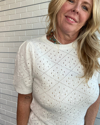 A smiling blonde woman wearing a white short-sleeve knit top with an eyelet diamond pattern, standing against a white brick wall.
