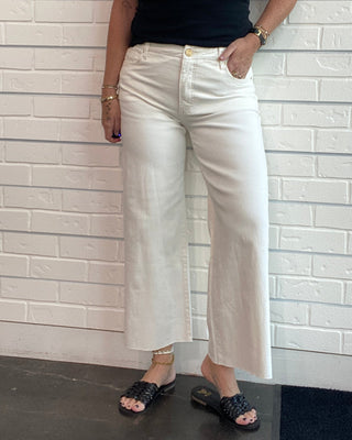 A woman wearing white wide-leg cropped pants, a black sleeveless top, and black slide sandals, posing against a white brick-patterned wall.