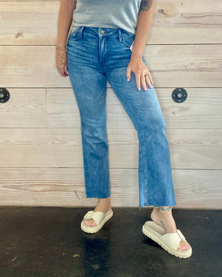 Person wearing a gray t-shirt, blue jeans, and white sandals standing against a wooden wall.