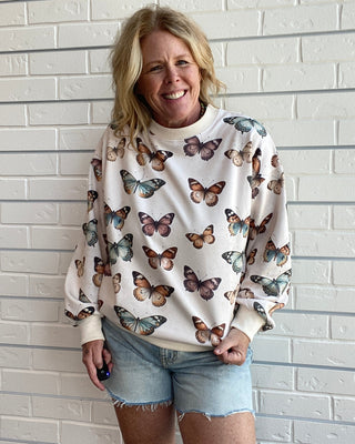 A woman smiling while wearing a cream-colored crewneck sweatshirt with an all-over butterfly print, paired with light-wash denim shorts, standing against a white brick wall.