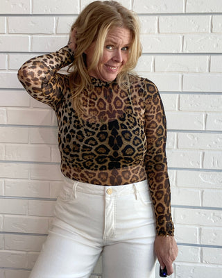A woman posing in a brown and black leopard print sheer long-sleeve mock neck top paired with white jeans, standing against a white brick wall.