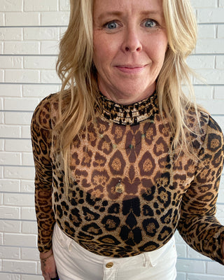 A woman wearing a sheer leopard-print long-sleeve mock neck top with a beaded collar, paired with white pants, standing against a white brick wall.