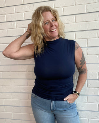 Woman wearing a navy sleeveless top and blue jeans standing against a light brick wall.