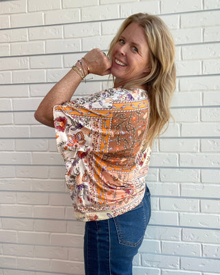 A woman with blonde hair wearing a short-sleeve patterned blouse and blue jeans, posing against a white brick wall.