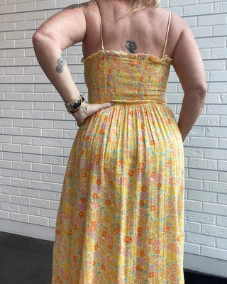 A woman viewed from behind wearing a yellow floral midi dress with thin spaghetti straps and a smocked bodice, standing against a white brick wall.