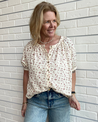 Woman wearing a floral blouse and jeans standing against a white brick wall.