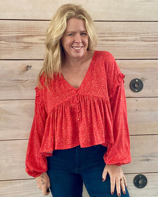 Woman wearing a red blouse and blue jeans standing against a wooden wall.
