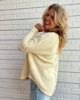 A woman with blonde hair wearing a cream ribbed knit long-sleeve sweater and a denim skirt, posing against a white brick wall.