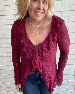 A smiling blonde woman wearing a long-sleeve burgundy lace top with ruffles and blue jeans, standing against a white brick wall.