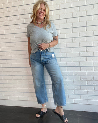 A woman modeling a light gray v-neck t-shirt and light-wash blue cropped jeans, standing against a white brick wall.