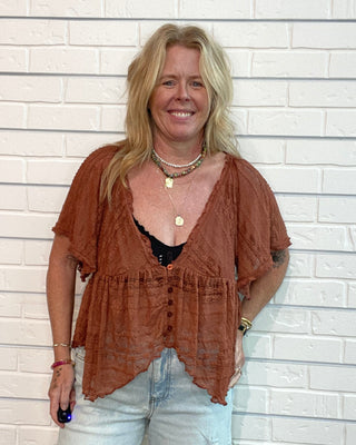 A woman with blonde hair stands against a white brick wall wearing a rust-brown short-sleeve textured top over a black bralette and light-wash distressed jeans.