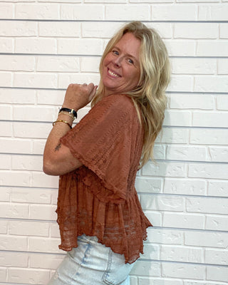 A woman with blonde hair wearing a rust-brown short-sleeve lace top and light-wash jeans, posing against a white brick wall.