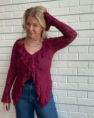A woman wearing a burgundy lace long-sleeve ruffled cardigan and blue jeans, posing against a white brick wall.