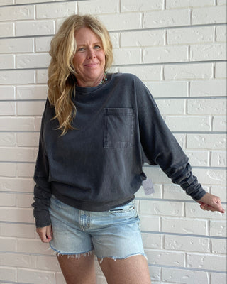 A woman with blonde hair stands against a white brick wall wearing a dark charcoal gray long-sleeve crewneck sweatshirt with a chest pocket and light-wash denim shorts.