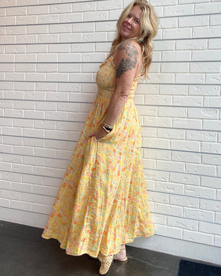 A woman with blonde hair and a visible tattoo on her upper arm is wearing a sleeveless yellow maxi dress with a small pink and green floral print, standing against a white painted brick wall.