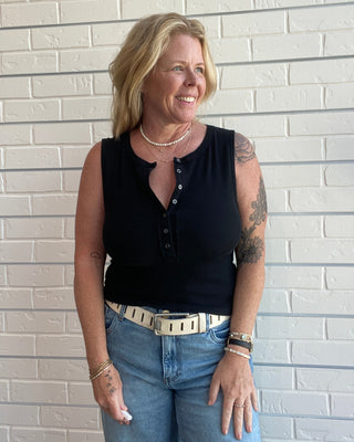 Woman wearing a black sleeveless top and blue jeans standing against a light brick wall.