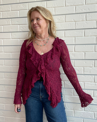 A woman wearing a burgundy lace long-sleeve ruffled top and blue jeans, standing against a white painted brick wall.