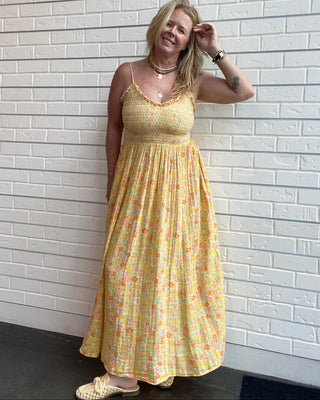 A woman wearing a yellow floral maxi dress with spaghetti straps, standing against a white brick wall.