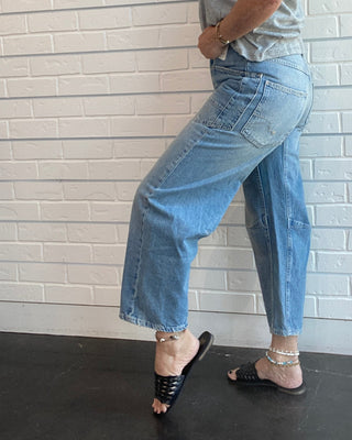 A woman posing in a light gray t-shirt and light blue cropped denim jeans against a white brick wall.