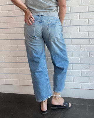 A woman is shown from behind wearing light-wash blue wide-leg jeans, a heather gray t-shirt, and black slide sandals, standing against a white brick wall.