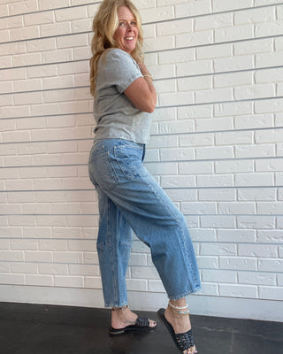A woman posing in a gray t-shirt and light blue cropped wide-leg jeans against a white brick wall.