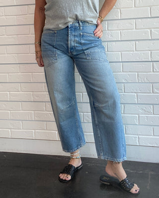 A woman wearing a light gray v-neck t-shirt, wide-leg cropped blue jeans, and black slide sandals, standing against a white brick wall.
