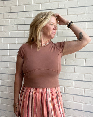 Woman wearing a brown top and pink striped pants against a white brick wall.