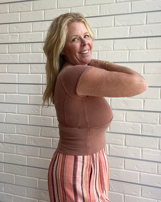 Woman wearing a brown top and striped pants against a light brick wall.