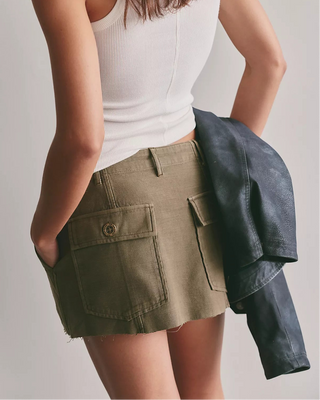 A woman shown from the back wearing a white ribbed tank top and an olive green cargo mini skirt, with a dark blue leather jacket draped over her arm, against a plain light gray background.
