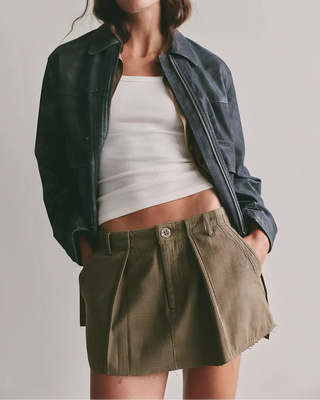 A woman modeling a dark blue leather jacket over a white ribbed crop top and an olive green pleated mini skirt, photographed against a neutral light gray studio background.