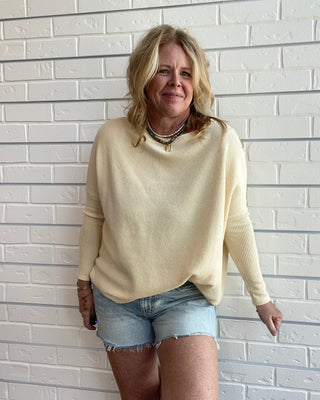 A woman with blonde hair wearing a cream-colored knit sweater and denim shorts, posing against a white brick wall.