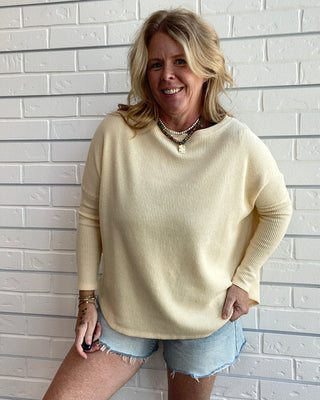 A woman wearing a pale cream long-sleeve knit sweater and light blue denim shorts, standing against a white painted brick wall.