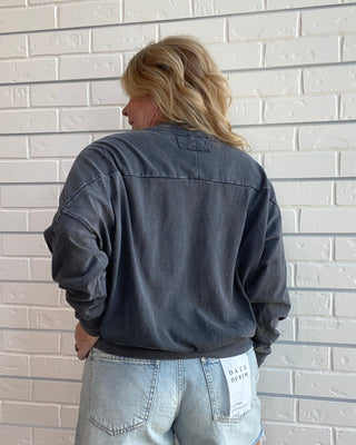 A woman viewed from behind wearing a washed charcoal gray long-sleeve button-up shirt and light-wash denim shorts, standing against a white brick wall.