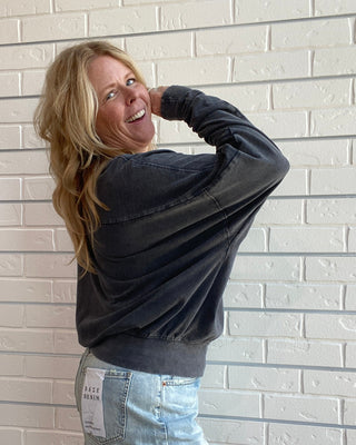 A woman posing in a dark washed long-sleeve sweatshirt and a light blue denim mini skirt against a white brick wall.