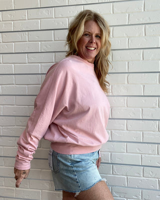 A smiling woman with blonde hair stands against a white brick wall, wearing a light pink long-sleeve crewneck sweatshirt and light blue denim shorts.