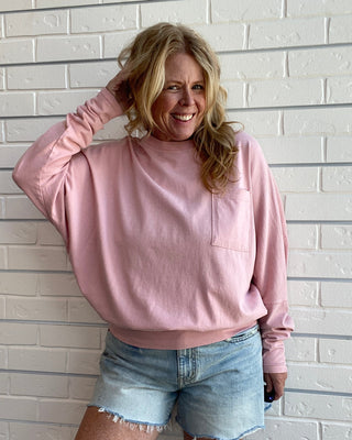 A woman wearing a dusty pink long-sleeve crewneck sweatshirt and light blue denim shorts, posing against a white brick wall.