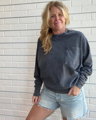 A woman with blonde hair wearing a dark gray washed long-sleeve crewneck sweatshirt with a chest pocket and light-wash denim shorts, standing against a white brick wall.