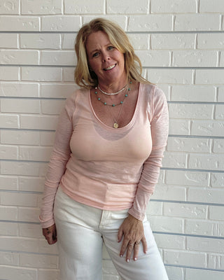 A smiling blonde woman wearing a light pink long-sleeve scoop-neck top and white pants, standing against a white painted brick wall.