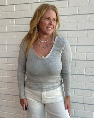 A woman with blonde hair wearing a light gray long-sleeve scoop neck top and white pants, standing against a white painted brick wall.