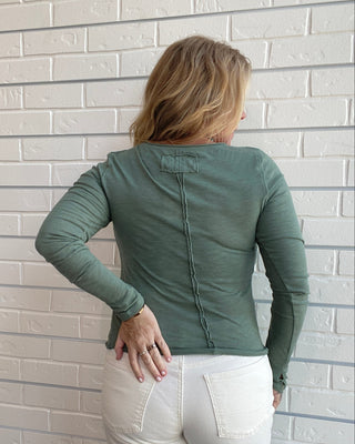 A woman viewed from behind wearing a sage green long-sleeve top and white pants, standing against a white brick wall.