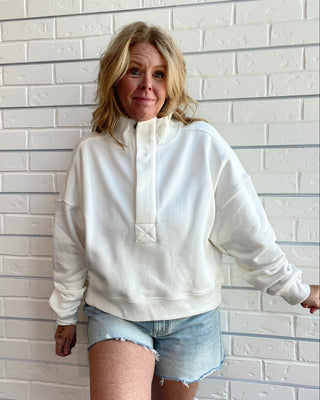 A woman wearing a cream-colored half-zip sweatshirt and light-wash denim shorts, posing against a white brick wall.