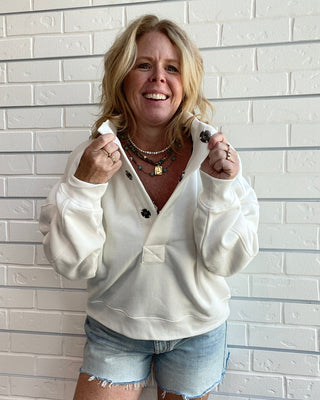 A smiling blonde woman stands against a white brick wall wearing a cream-colored knit zip-up sweater and light-wash denim shorts.