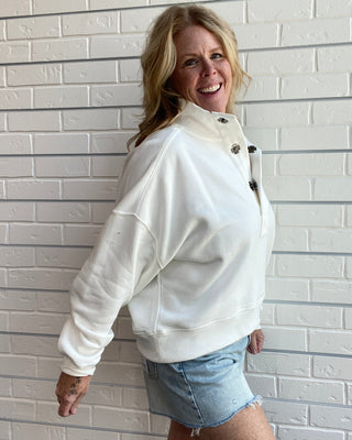 A woman smiling and posing in a white mock-neck sweatshirt with dark buttons and light blue denim shorts, standing against a white painted brick wall.