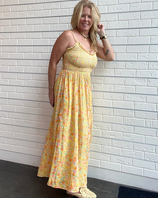 A woman wearing a yellow floral maxi dress with spaghetti straps and a smocked bodice, standing against a white brick wall.