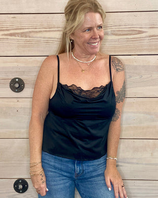 Woman wearing a black top and blue jeans standing against a wooden wall.