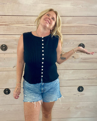 Woman wearing a navy sleeveless top with white buttons and denim shorts against a wooden wall.