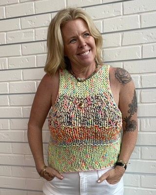 Woman wearing a colorful sleeveless top and white shorts standing against a light brick wall.