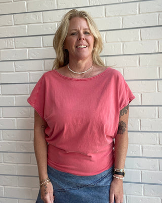 Woman wearing a pink shirt and blue jeans standing against a white brick wall.