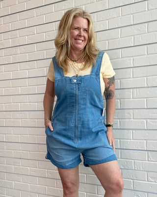 Woman wearing a denim romper standing against a light brick wall.