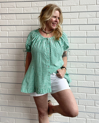Woman wearing a green checkered top and white shorts standing against a light brick wall.